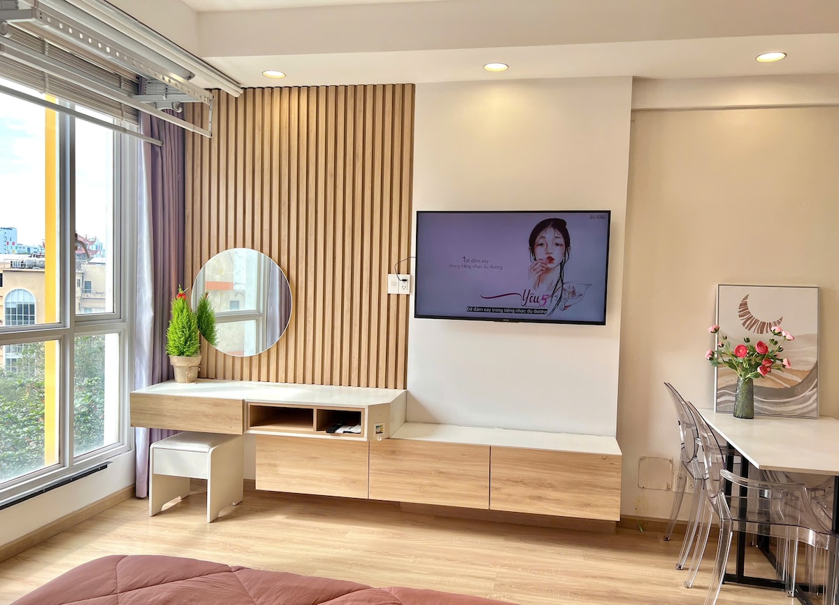 A modern studio space is displayed, featuring a wall-mounted television above a light wood cabinetry. A round mirror and a potted green plant add natural elements. A glass dining table with transparent chairs is positioned near a large window, allowing ample natural light.