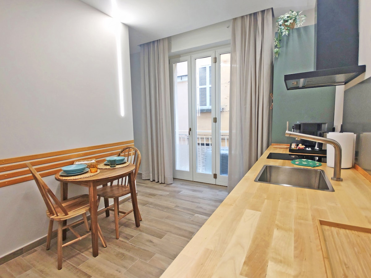 A modern kitchen area features a wooden countertop and a sink, with a view through glass doors leading to a small balcony. A round table set for two with wooden chairs occupies the corner, complemented by soft curtain drapes that allow natural light to brighten the space.
