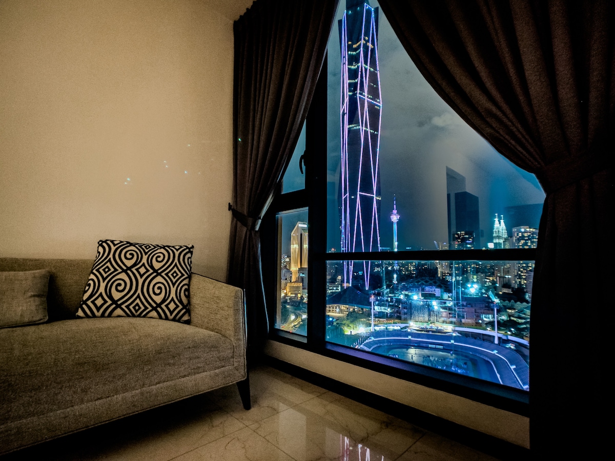 A corner of the living area showcases a comfortable sofa adorned with a geometric patterned cushion. A large window offers a stunning view of the illuminated city skyline, featuring the KL Tower and modern high-rise buildings amidst a night sky.