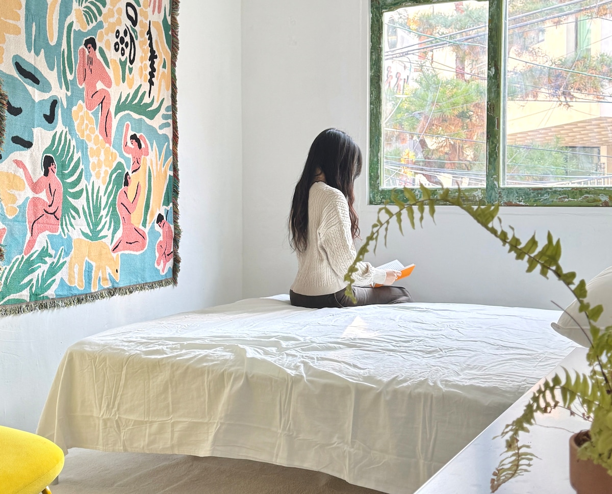 A spacious bed is centrally located in the bright room, featuring a colorful wall tapestry depicting abstract figures. Natural light filters through large green-framed windows, illuminating the interior. A cozy corner is created by a green fern and a minimalistic furniture piece nearby.