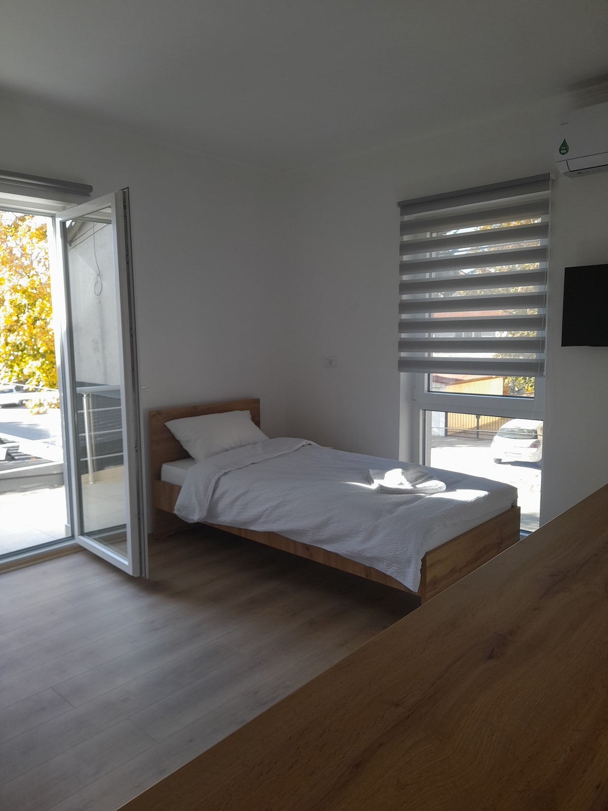A simple bedroom features a spacious bed with a white blanket. Natural light flows in through the partially open door leading to a small balcony, while modern window treatments are drawn over a window. A wall-mounted television is visible.