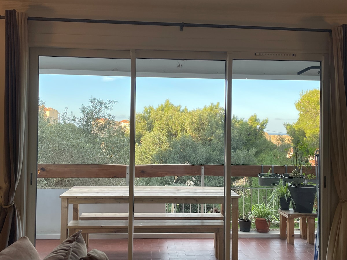 A spacious balcony is visible through large glass doors, featuring a wooden table surrounded by chairs. Various potted plants are positioned along the railing, with greenery providing a serene backdrop. Soft natural light filters into the living space, enhancing the welcoming atmosphere.