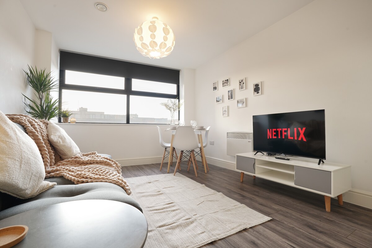 A well-lit living area features a comfortable sofa with textured throws, a coffee table, and a Smart TV displaying Netflix. A dining table for two sits nearby, with light streaming in from large windows covered by sleek blinds. Wall-mounted photographs add a personal touch.