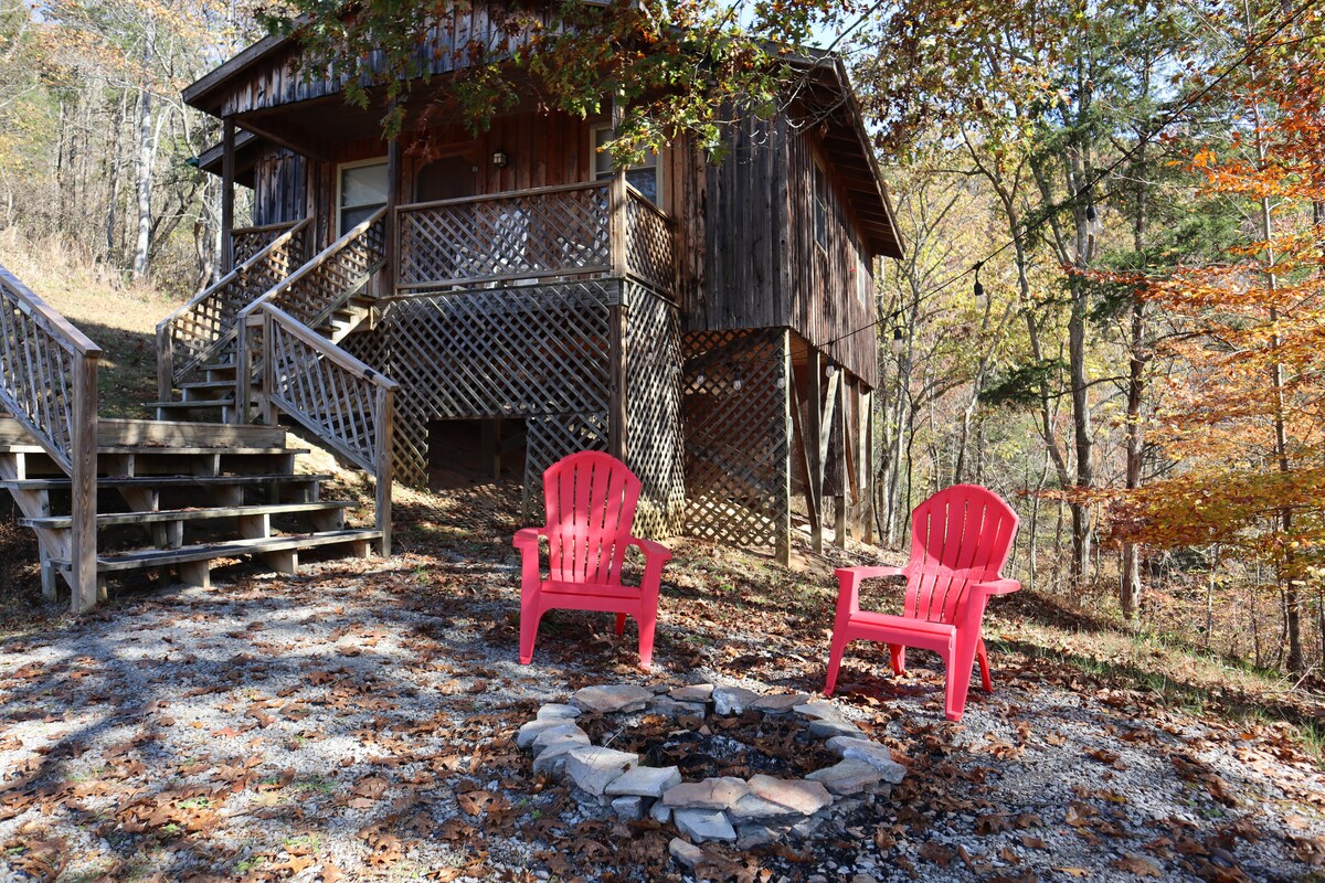 A rustic wooden cottage is elevated on stilts, surrounded by trees with autumn foliage. Two bright pink Adirondack chairs are positioned around a circular stone fire pit, inviting relaxation outdoors. Steps lead up to the cottage's entrance from the gravel pathway.