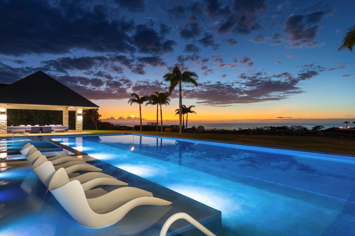 An inviting pool area is illuminated by soft lighting, with lounge chairs positioned along the water's edge. The tranquil pool reflects the vibrant colors of a sunset over the Pacific Ocean, while silhouettes of palm trees stand against the colorful sky.