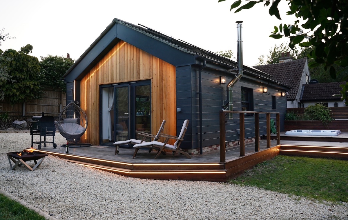 The exterior of The Annexe is highlighted by a modern wooden facade alongside dark siding. A spacious deck features two lounge chairs and a hanging chair, with soft lighting enhancing the area. The hot tub is positioned to the right, surrounded by a well-kept lawn.