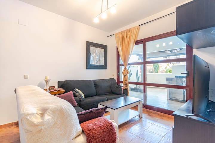 סלון גדול עם ספה אפורה ועוד שולחן קפה דו-צבעוני, ריהוט כהה, טלוויזיה, מיטת קנופי ומסך, פינת אוכל עם שולחן וכיסאות גבוהים, מדף עץ, חלונות עם יציאה למרפסת מקורה ורצפת קרמיקה בגוון חם.
