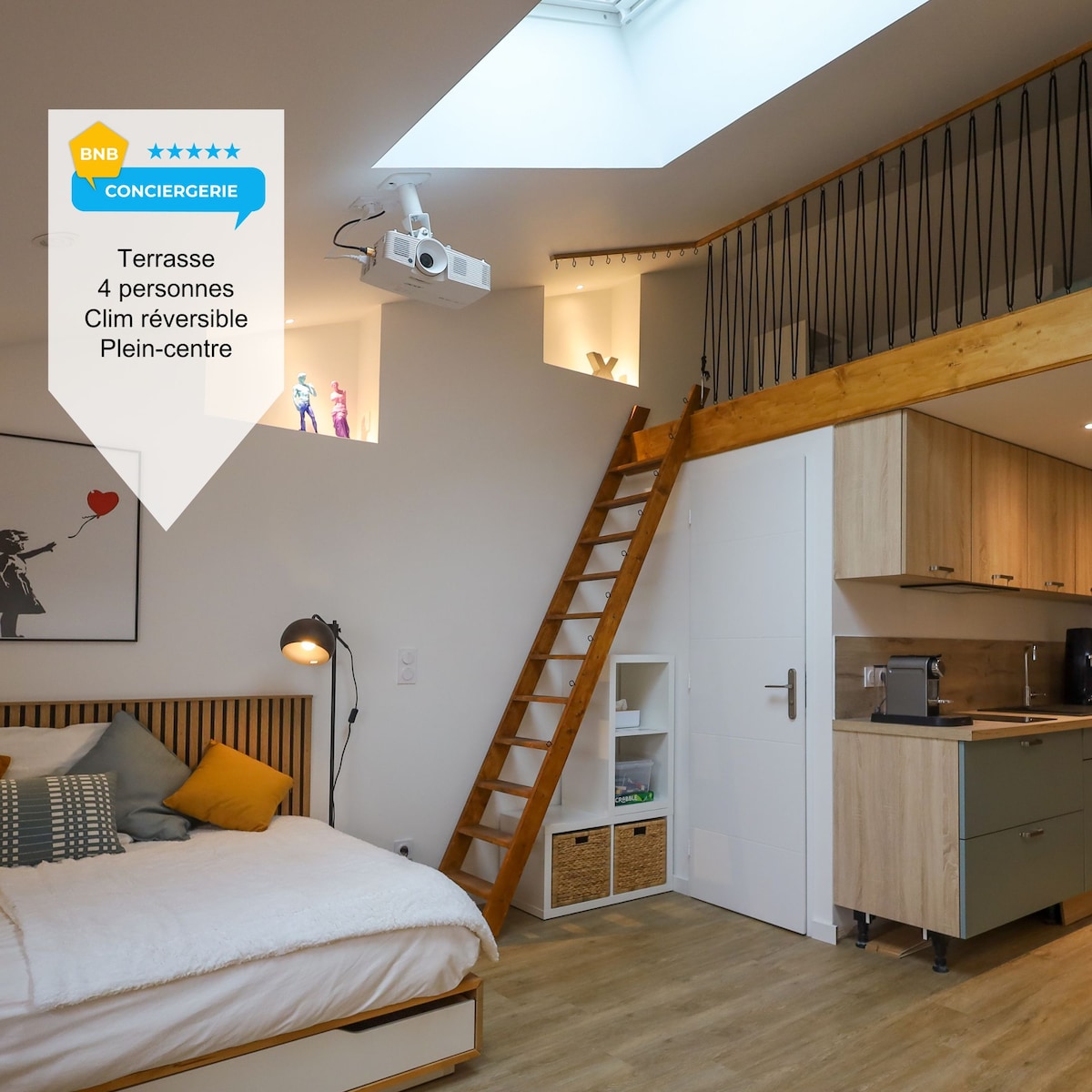 A spacious room is illuminated by natural light from a skylight above. A cozy sleeping area features a comfortable bed, while a wooden ladder leads to a mezzanine. A modern kitchen area is visible in the background, providing convenient amenities for guests.