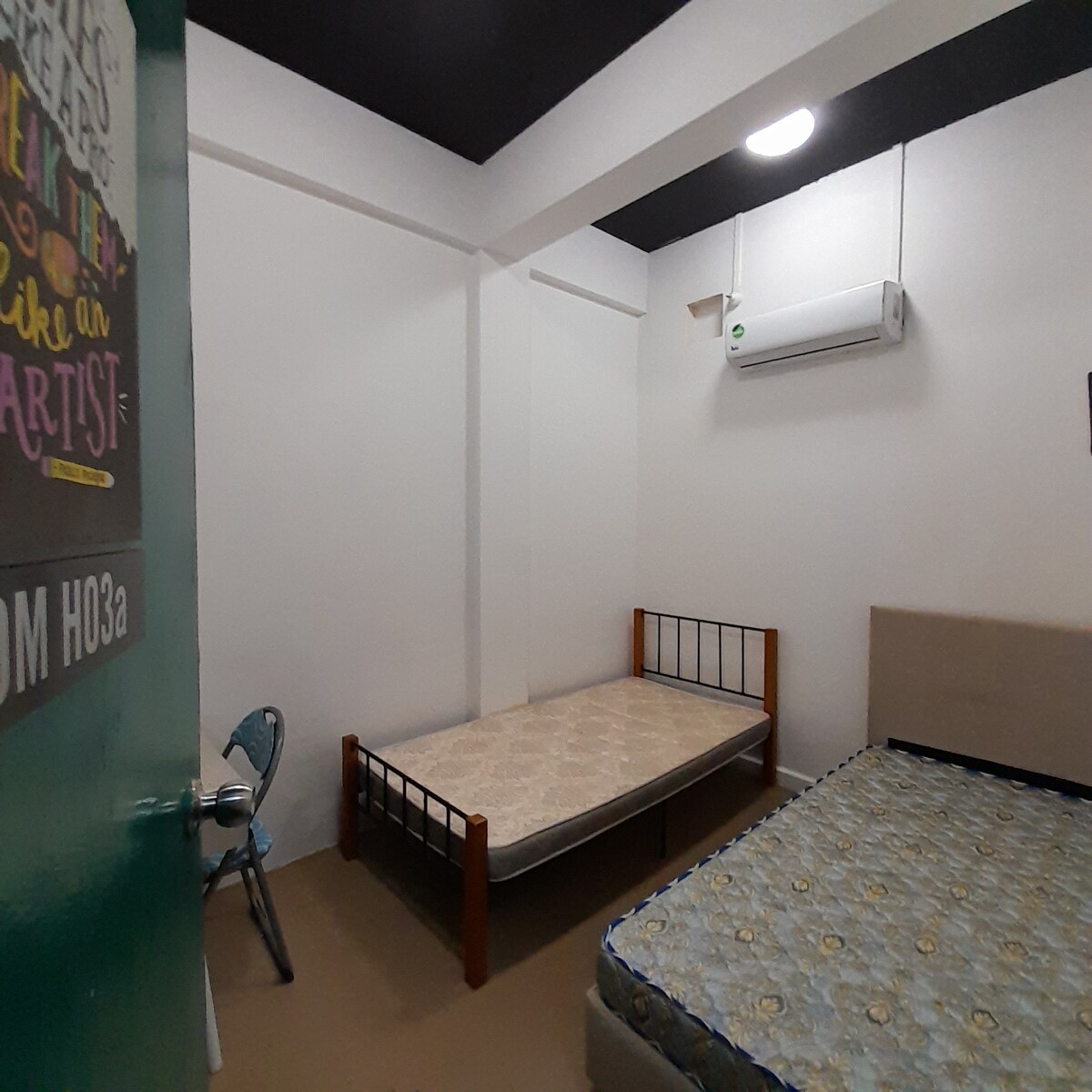 A simple room features two single beds, one of which is fully made and the other left unmade. A metal chair is positioned nearby. The space is well-lit by overhead lighting, and an air conditioning unit is mounted on the wall.