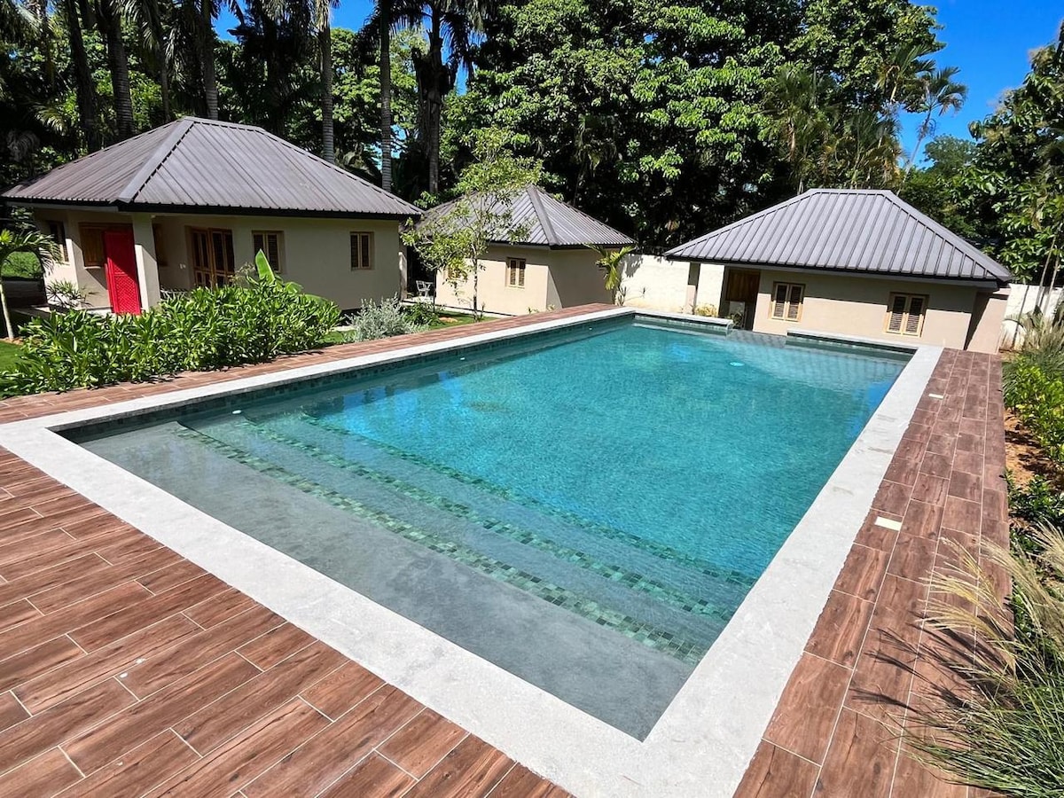 A serene rectangular swimming pool is surrounded by a wooden deck, providing a calm atmosphere. Lush tropical foliage frames the pool area, and two bungalows with neutral walls and dark roofs are visible in the background. Clear water reflects the greenery overhead.