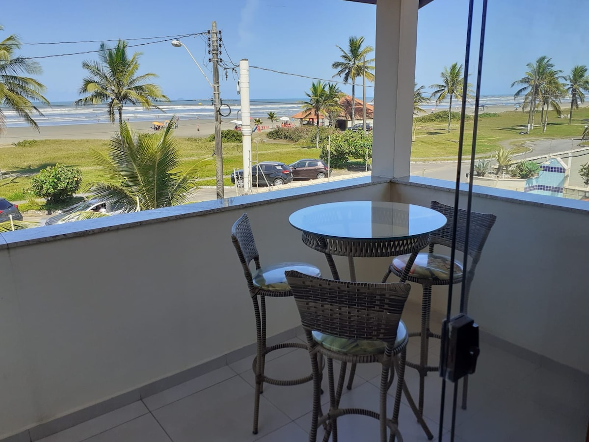 A balcony area features a round table surrounded by four woven chairs. A view of the beach and palm trees is visible through large glass doors, inviting natural light and a sense of openness.