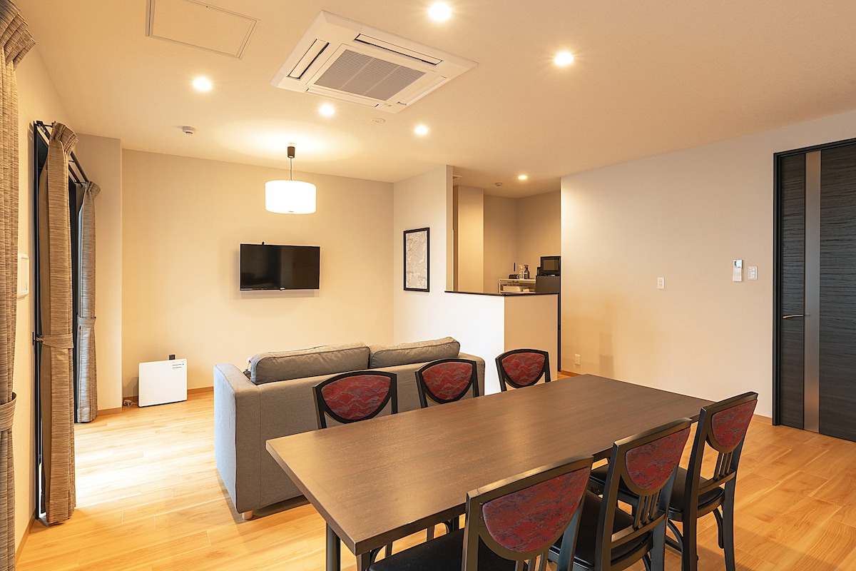 The spacious living-dining area features a comfortable sofa and a large dining table surrounded by multiple chairs. A flat-screen TV is mounted on the wall, and soft lighting enhances the warm ambiance. The kitchen area is visible in the background.