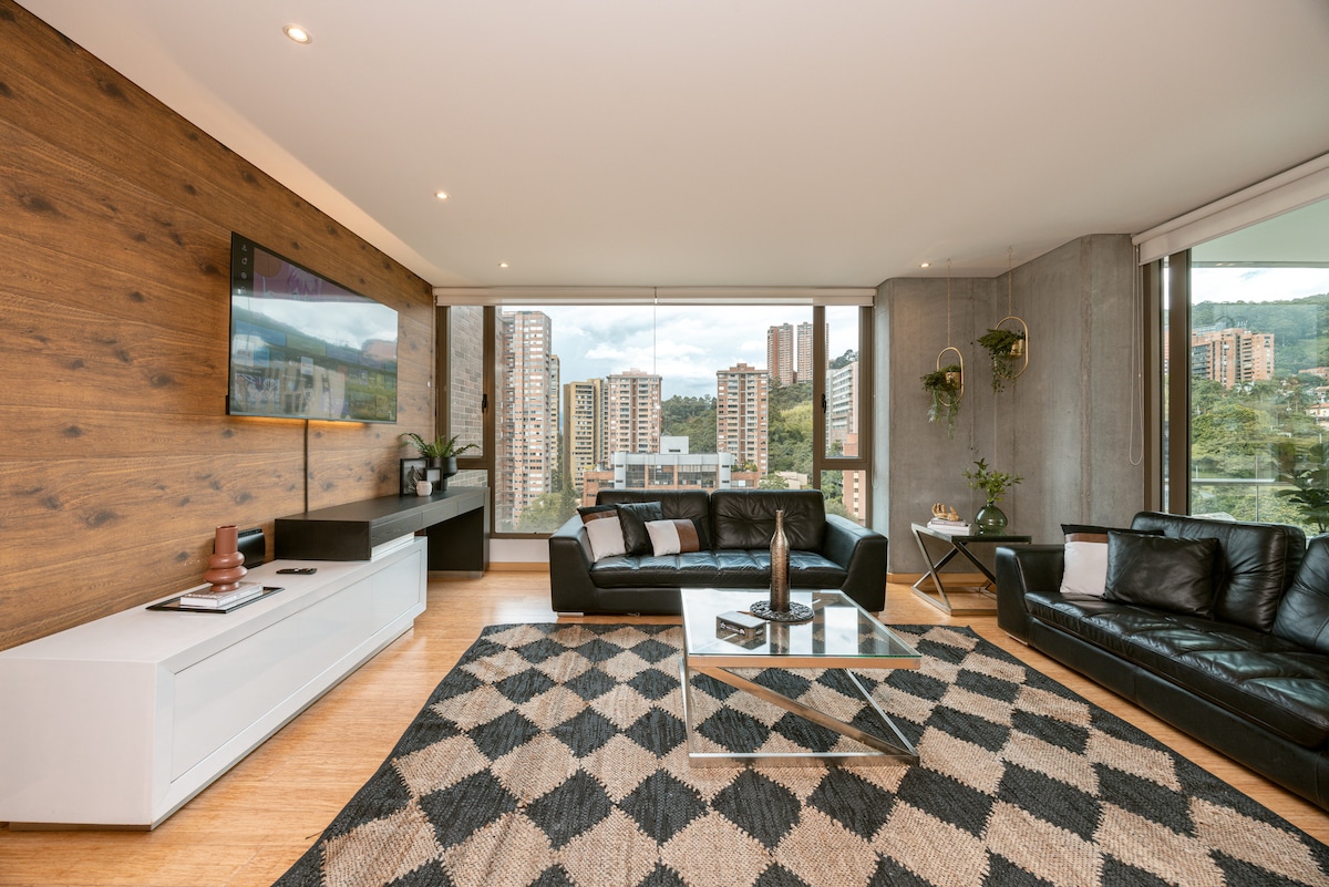 The spacious living area features modern furnishings, including two black sofas and a sleek coffee table. A large television is mounted on a wood-paneled wall, while floor-to-ceiling windows provide views of Medellín's skyline. A patterned rug adds texture to the wooden floor.