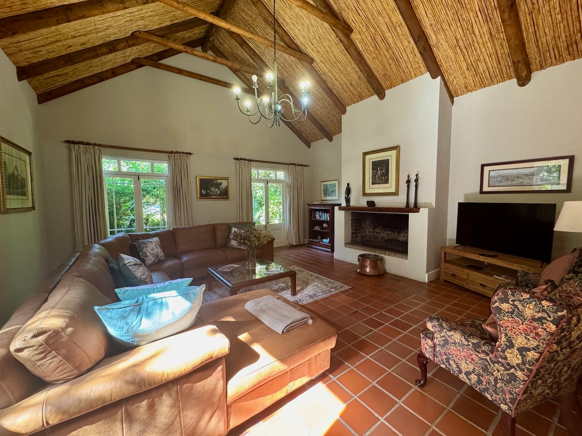 A spacious lounge area features a large sectional sofa with decorative pillows, a cozy armchair, and a wooden coffee table. Natural light streams through multiple windows, illuminating the rustic ceiling with wooden beams. A fireplace and a Smart TV are included along one wall.