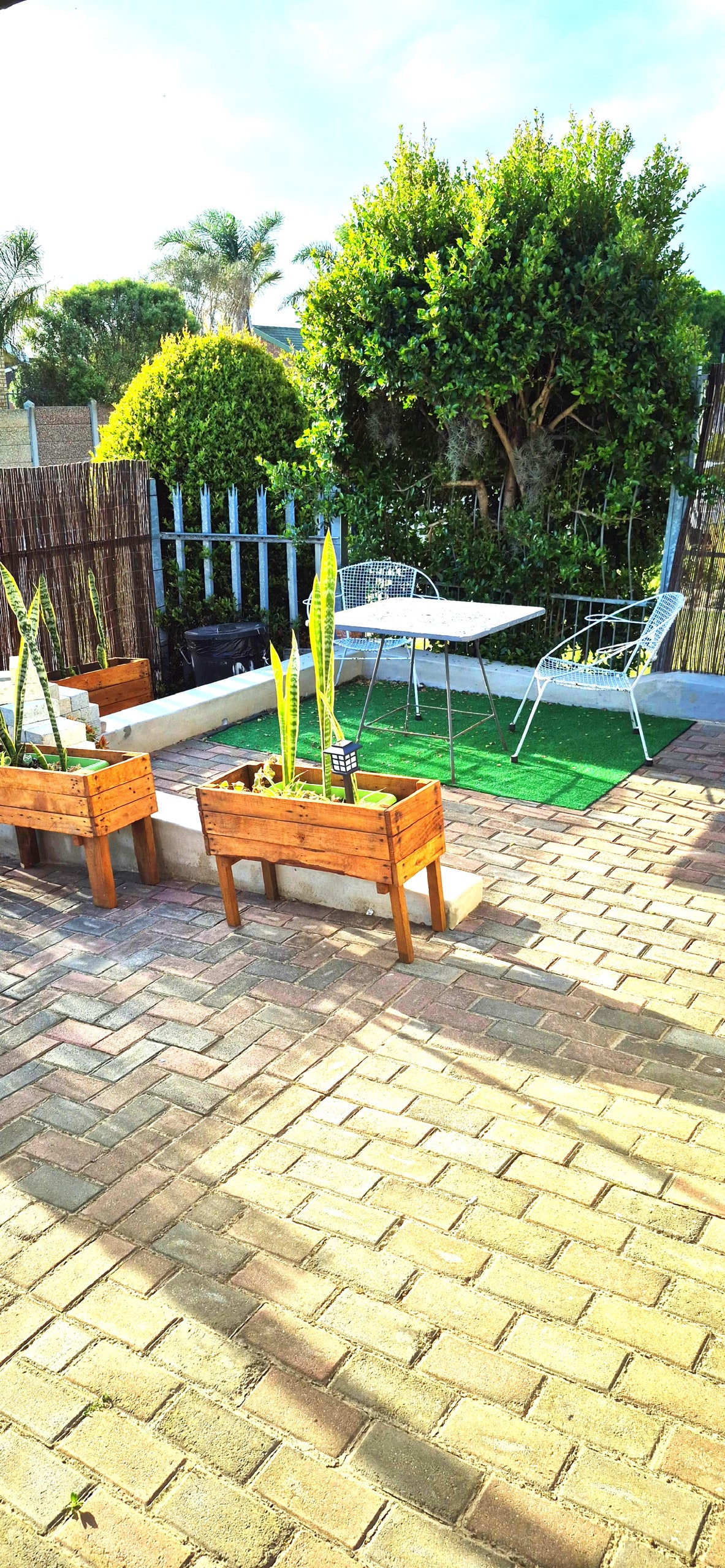 An outdoor seating area features a small table surrounded by two metal chairs. Wooden planter boxes with greenery are positioned on a paved surface, which is bordered by low hedges and a fence. The space is illuminated by natural light.
