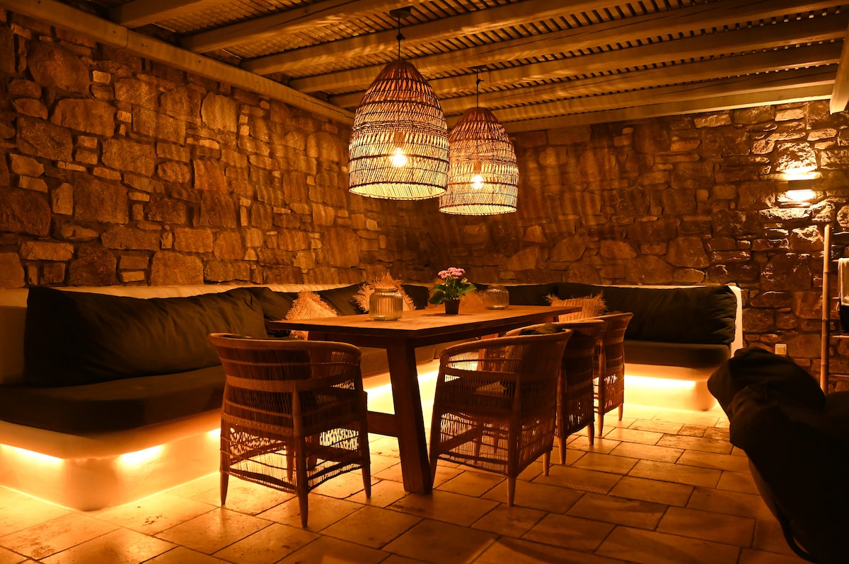 An inviting outdoor dining area is illuminated by warm lighting, featuring a large wooden table surrounded by wicker chairs. Soft seating along the stone wall provides a cozy atmosphere, while decorative pendant lights hang above, enhancing the intimate setting.