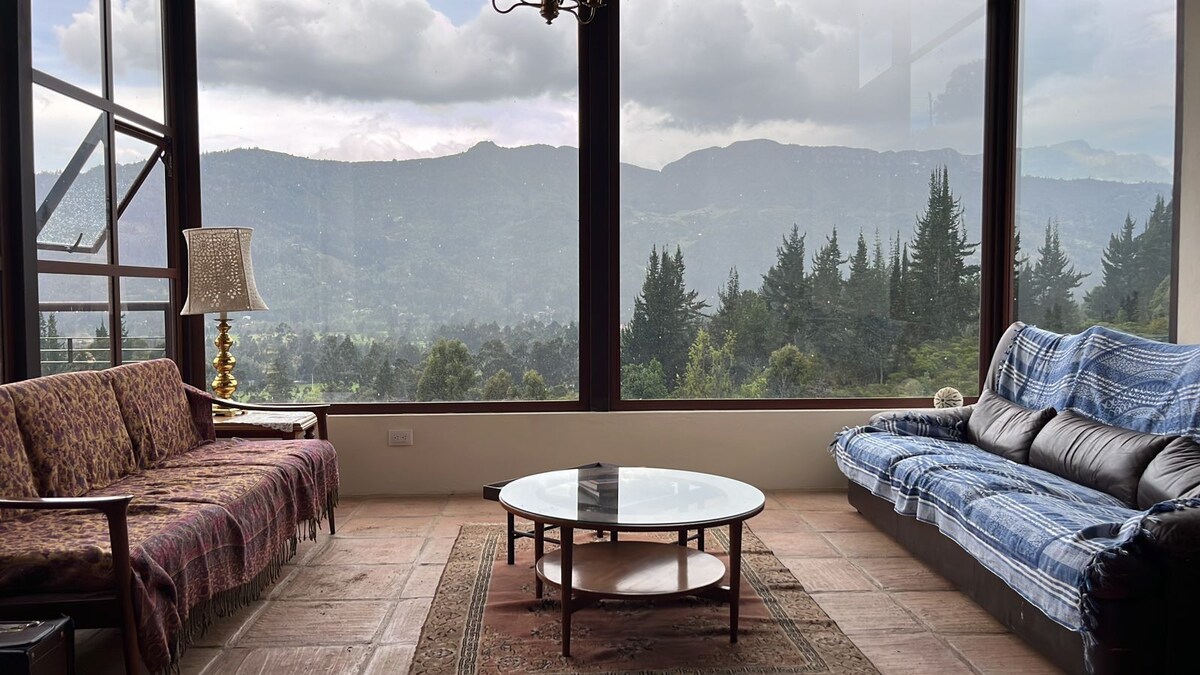 A spacious living area features large windows offering unobstructed views of the surrounding mountains. Two sofas provide seating, while a round coffee table sits on a decorative rug in the center. Natural light fills the space, enhancing the serene ambiance.