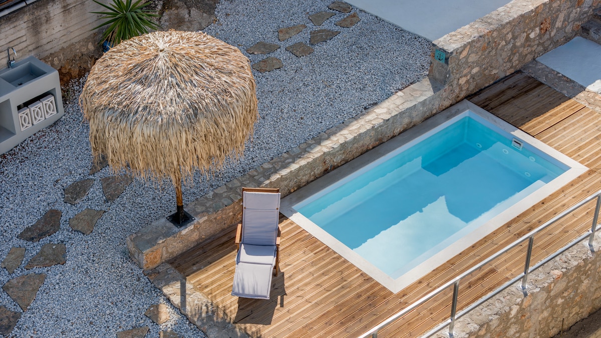 A private pool is surrounded by wooden decking and gravel, with a thatched umbrella providing shade. A single lounge chair is positioned beside the pool, creating a serene setting for relaxation. Natural stone elements are integrated into the surrounding landscape.