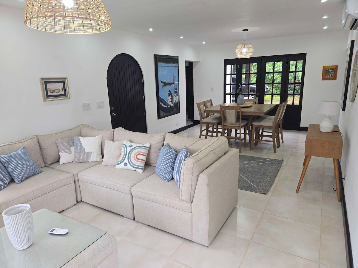 A light-filled living area features a spacious sectional sofa adorned with decorative cushions. A dining table with several chairs is visible in the background, illuminated by natural light streaming through large glass doors. Artwork and a stylish lamp complete the inviting setting.