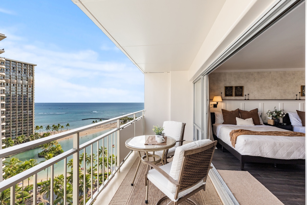 A private balcony lanai is showcased, featuring a table with two chairs overlooking an ocean view. The bedroom area is visible through large glass sliding doors, where a cozy bed is adorned with neutral-toned bedding. Lush palm trees can be seen framing the picturesque scenery.