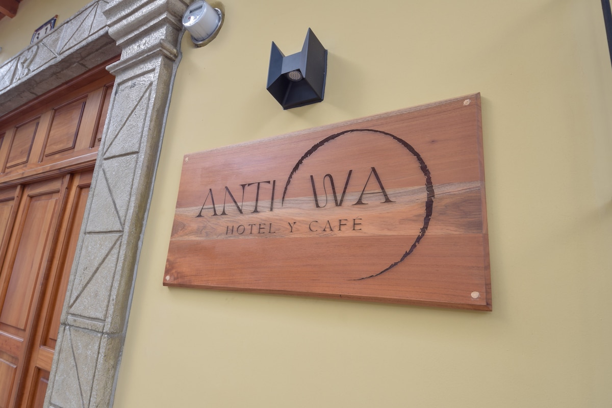 The exterior signage of the hotel presents the name 'ANTIGUA HOTEL Y CAFÉ' on a wooden plaque. The sign is attached to a warm yellow wall, complemented by a wall-mounted light fixture. The entrance features intricate stonework and a wooden door, inviting guests.