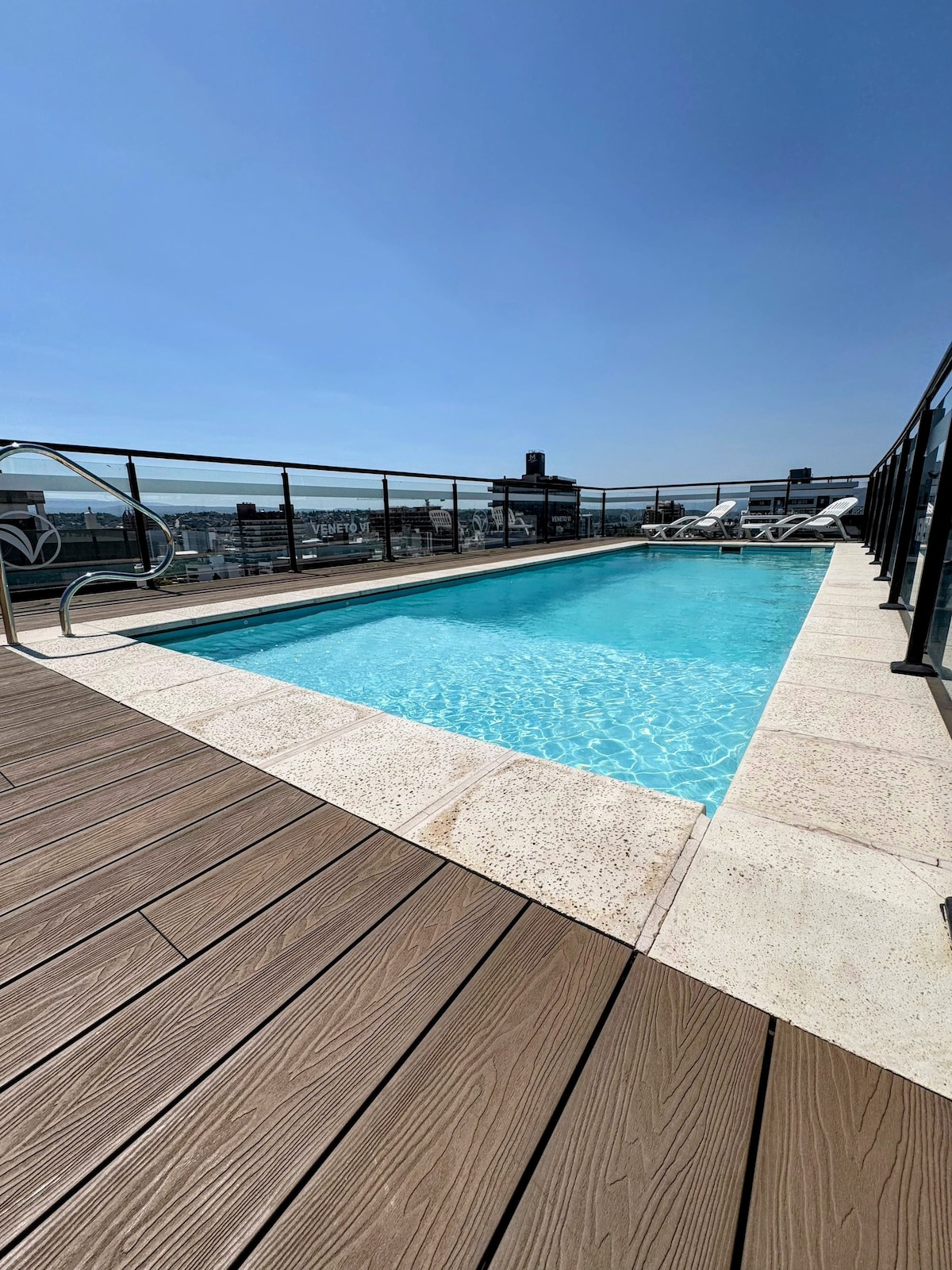 The rooftop area features a rectangular outdoor pool surrounded by wooden decking. Sun loungers are positioned nearby for relaxation. A clear sky enhances the spacious atmosphere, offering an unobstructed view of the surroundings.