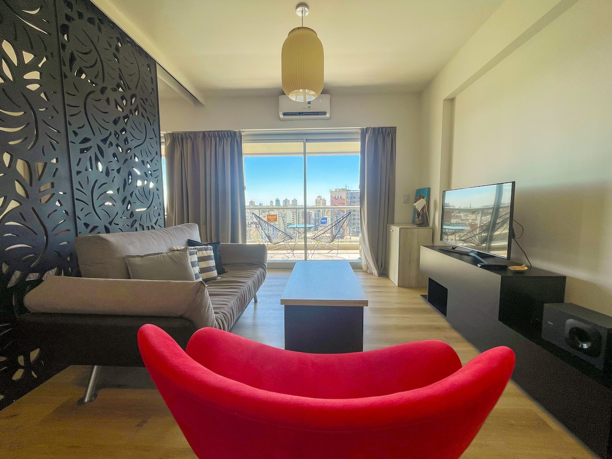 A modern living room features a spacious sofa, a vibrant red accent chair, and a wooden coffee table. Natural light streams through large windows, showcasing views of the cityscape. A smart TV is positioned on a low black entertainment unit, enhancing the contemporary style.