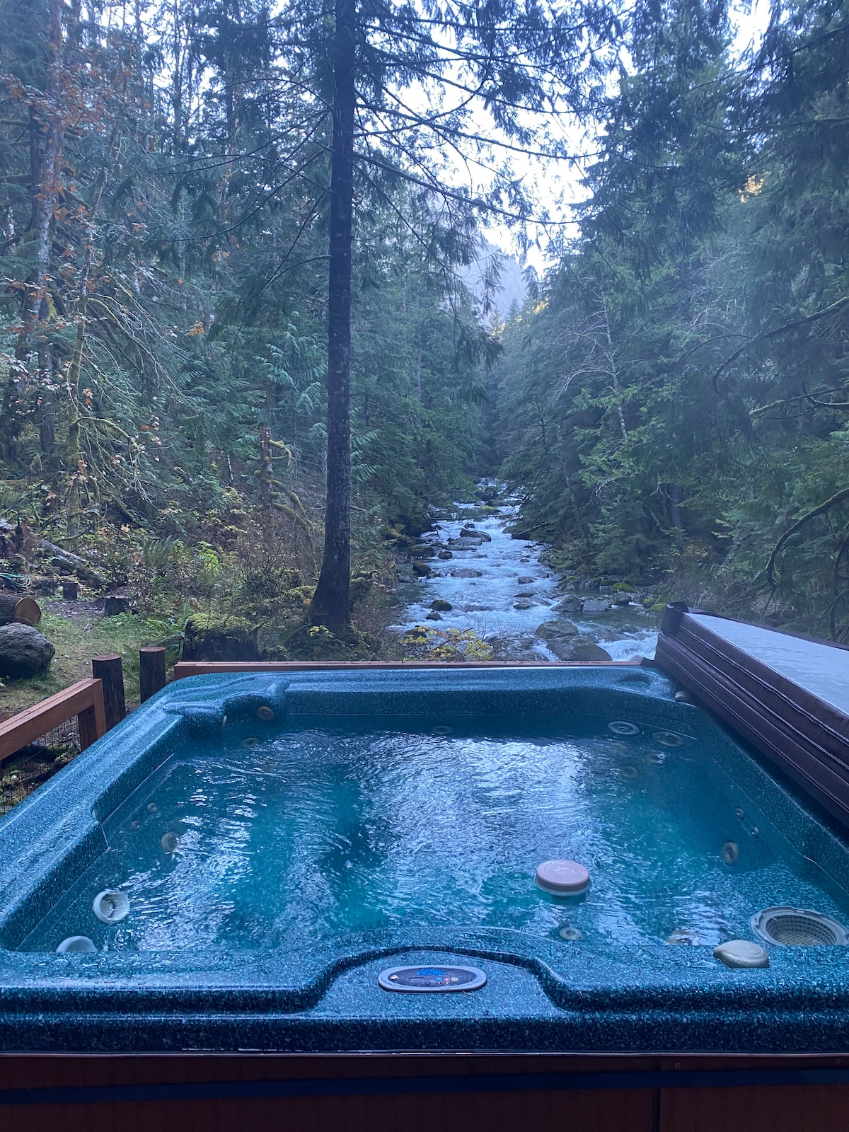 A hot tub is situated on a deck, surrounded by tall trees and a view of a flowing stream below. The water appears clear with bubbles visible, creating a serene outdoor space for relaxation.
