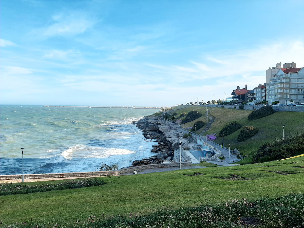 A scenic view captures the coastline of Playa Chica, showcasing undulating waves against rocky formations. Lush green hills lead to well-maintained pathways, with a mix of buildings and residential areas visible in the background. The sky is clear, contributing to a bright and airy atmosphere.