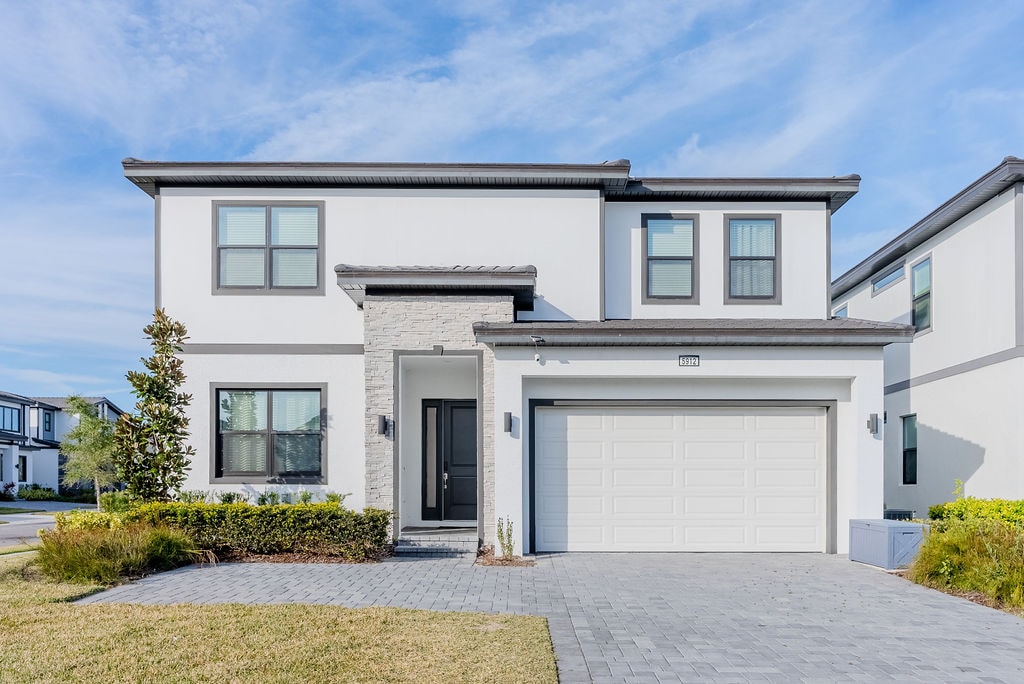 The exterior of a two-story home presents a modern design, featuring a light-colored facade with contrasting elements. A spacious driveway leads to a garage, while manicured landscaping adds a touch of greenery. Large windows allow for ample natural light, enhancing the inviting appearance.
