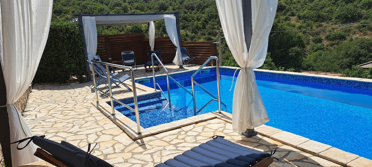 A refreshing swimming pool is surrounded by a stone patio, featuring a partial shade structure with draped fabric. Lounge chairs are positioned nearby, providing a comfortable space for relaxation, while lush greenery borders the area, enhancing the natural setting.