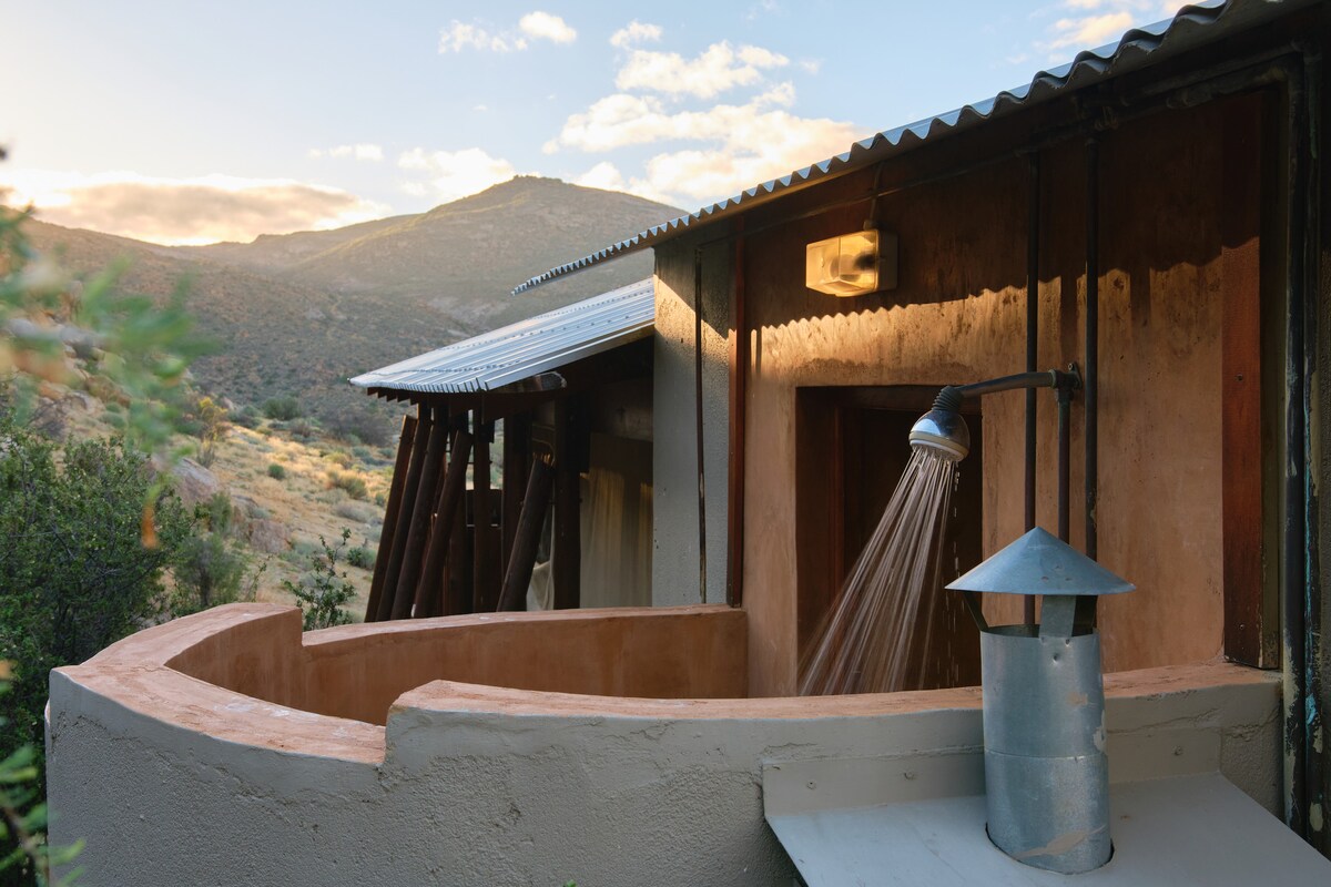 An outdoor shower is positioned against a rustic wall, with a gentle stream of water illuminated by ambient light. The surrounding landscape features rolling hills in the background, complementing the earthy tones of the structure and creating a natural and serene environment.