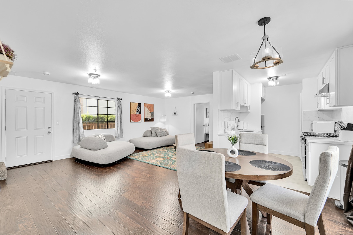 A bright and spacious living area features two modern sofas, a round dining table, and a well-equipped kitchen. Natural light filters through a large window, illuminating the room's neutral color palette and wood flooring. Decorative wall art adds subtle visual interest to the space.