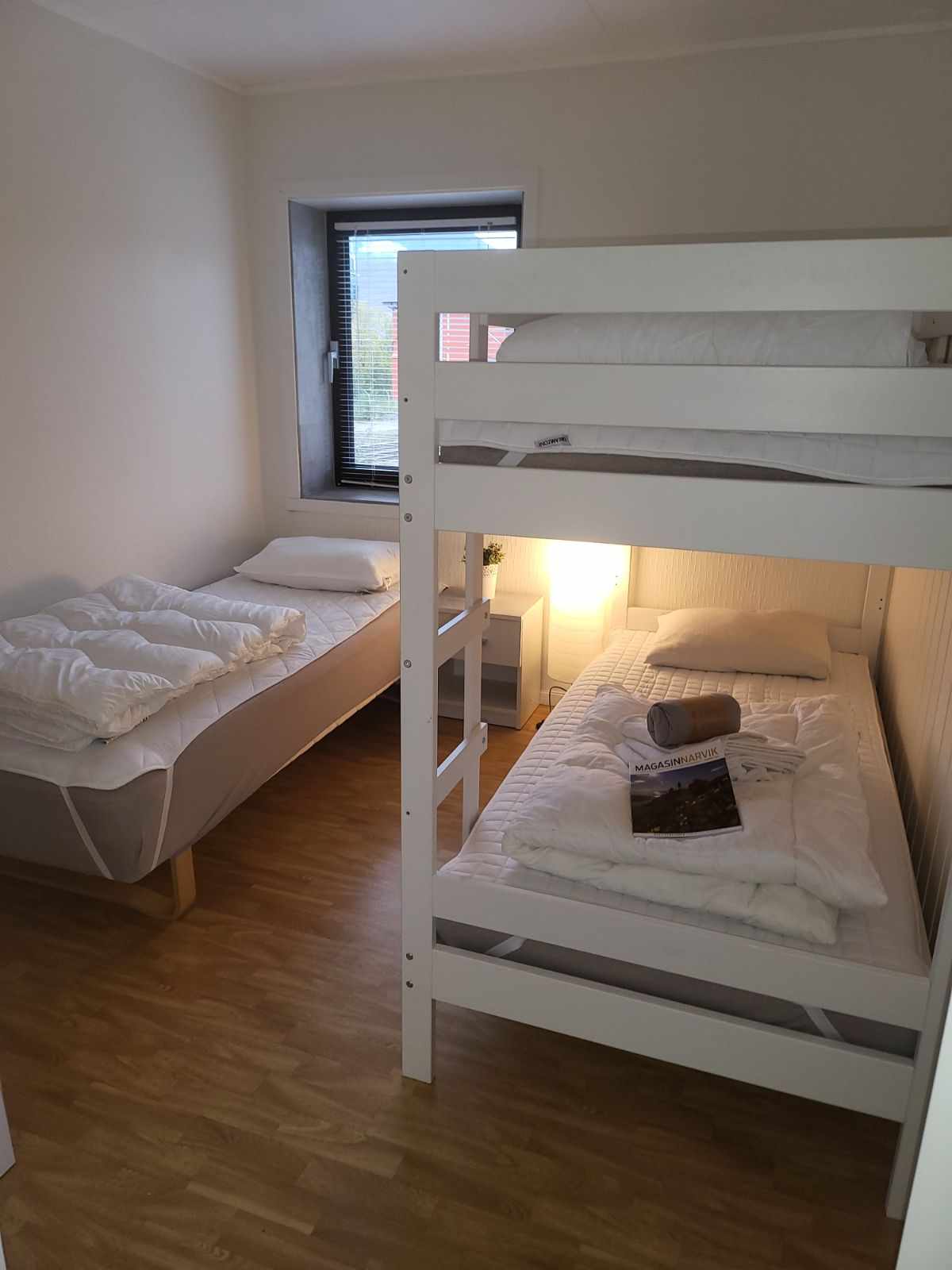 A well-lit bedroom is presented, featuring a bunk bed with a spacious single bed underneath and a mattress on the upper level. A bedside lamp adds soft illumination. Crisp bedding is neatly arranged on both beds, providing a clean and inviting space.