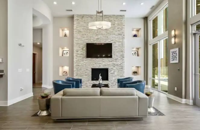 A modern lobby area features a light-colored seating arrangement facing a contemporary stone fireplace. High ceilings enhance the spaciousness, while large windows allow natural light to illuminate the space. Blue accent chairs and decorative wall niches add subtle pops of color.