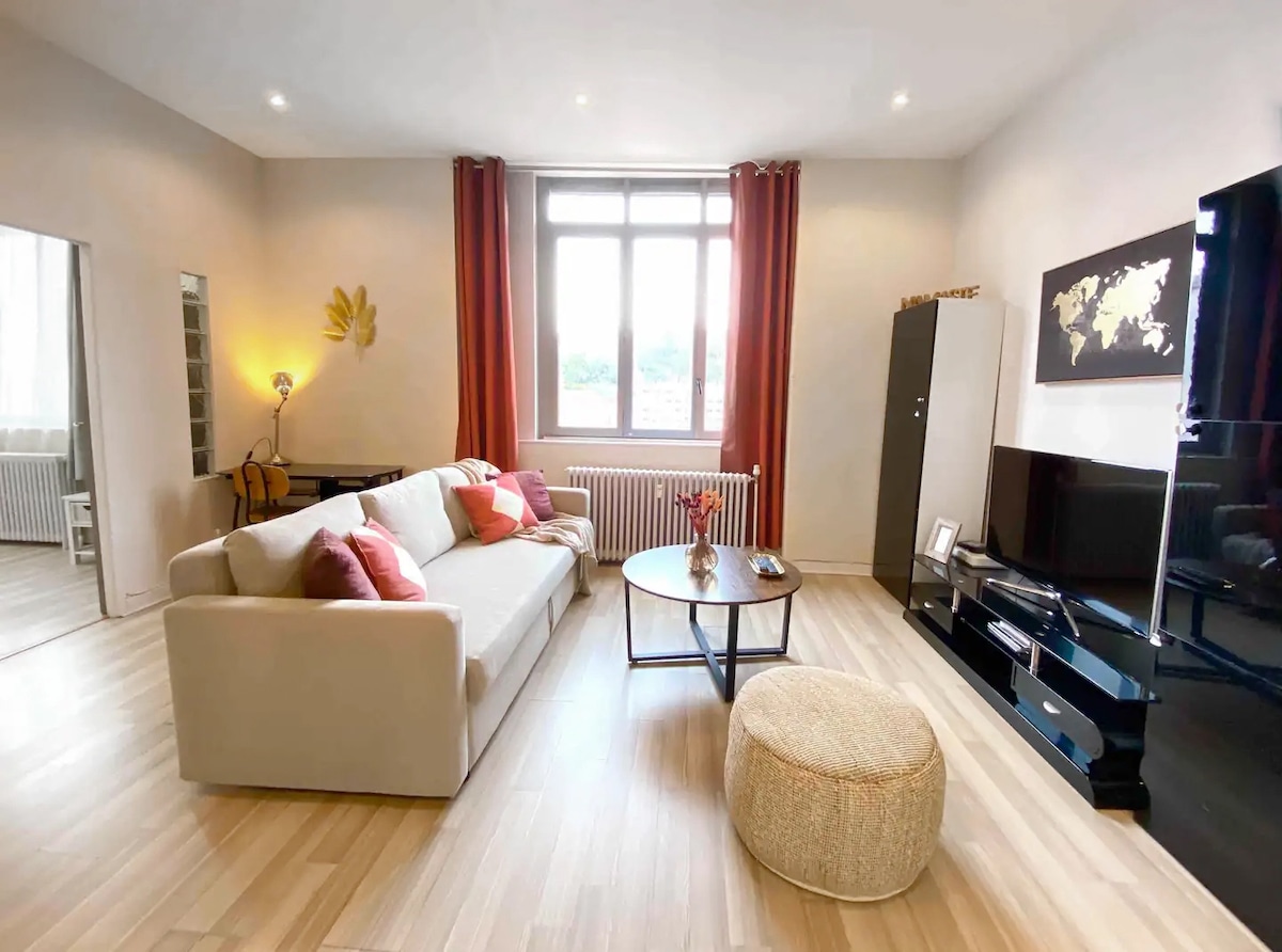 The spacious living area features a light-colored sofa adorned with decorative pillows, complemented by a round ottoman. A large window provides natural light, and a flat-screen television is mounted on the wall. Subtle decor accents the modern furnishings and warm wood floors.