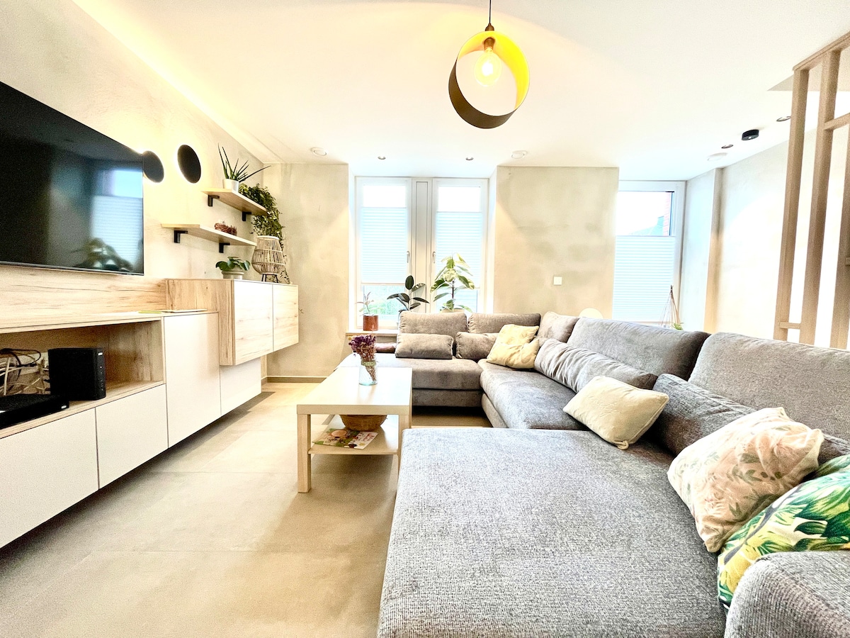 A modern living room features a large, grey sectional sofa adorned with decorative cushions. A light wood coffee table sits in front, complemented by a wall-mounted television. Natural light enters through large windows, creating a bright and inviting space, while indoor plants add a touch of greenery.