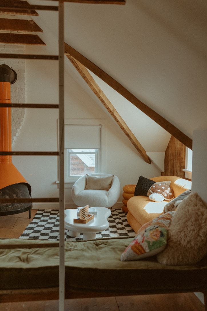 A cozy living area showcases a mix of textures with a light gray armchair, a bright orange couch, and a patterned black and white rug. Large windows allow natural light to illuminate the space, complemented by the warm tones of exposed wooden beams.