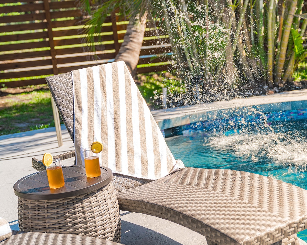 A comfortable lounge chair is positioned beside a sparkling pool, draped with a striped towel. Two refreshing beverages with lemon slices rest on a nearby table, while water splashes gently in the background, surrounded by greenery and a privacy fence.