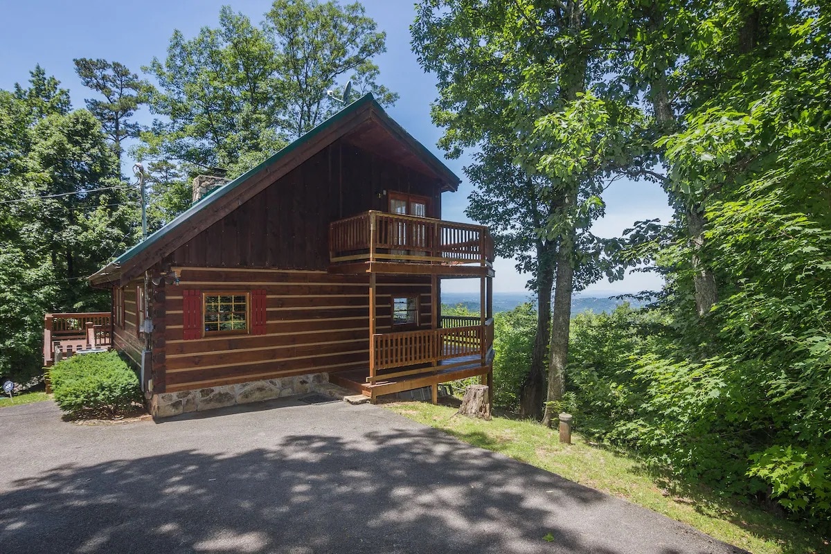 The exterior of a rustic log cabin is displayed, featuring a green roof and wooden balcony. The cabin is surrounded by lush trees and offers a glimpse of mountain views. A paved driveway leads up to the cabin, creating an inviting entrance.
