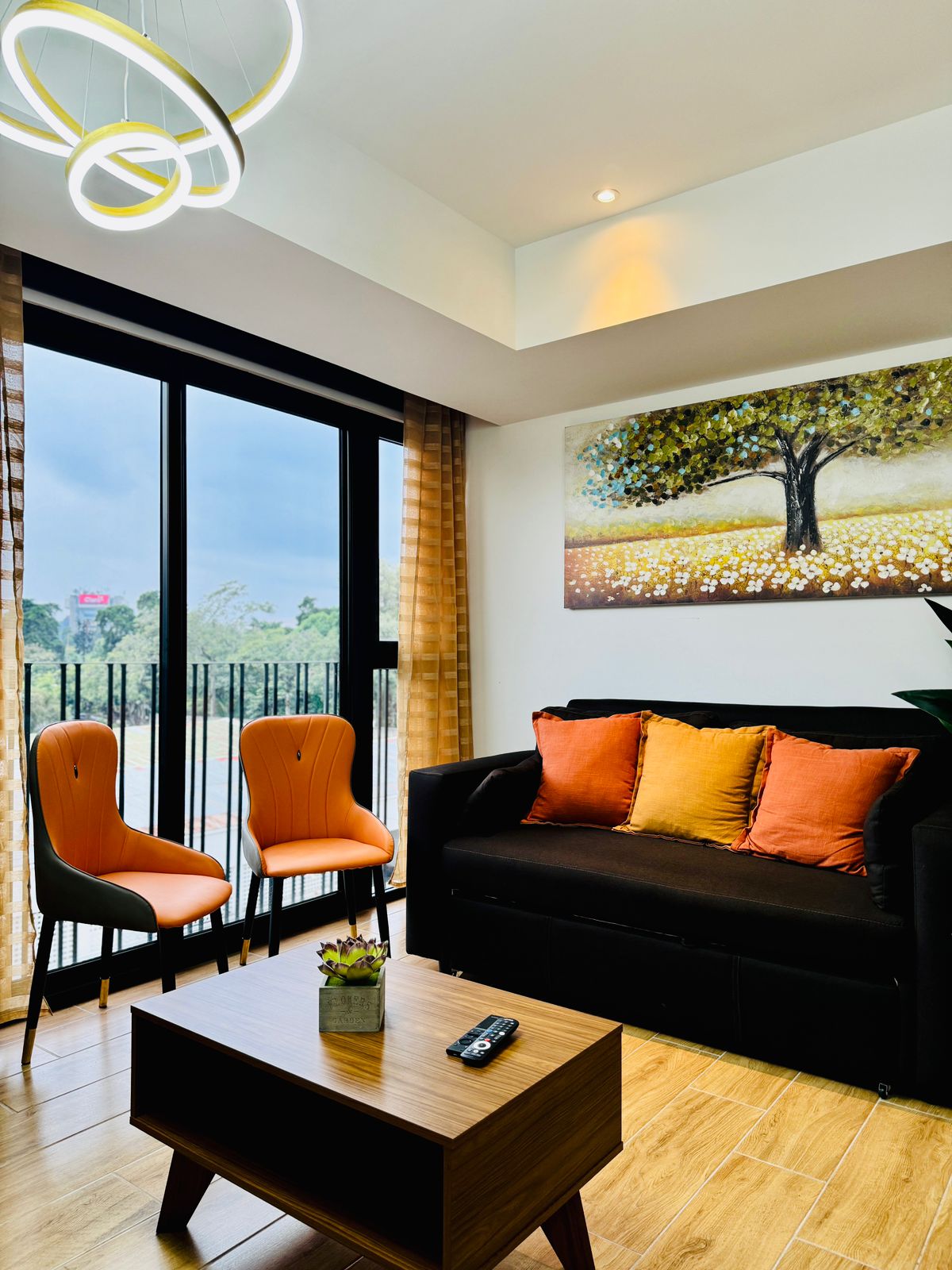 A cozy living area is presented with a dark sofa adorned with bright orange cushions. Two orange chairs complement the seating, while a wooden coffee table sits in the center. A wall art piece features a tree and a field of flowers, and natural light enters through large glass doors.