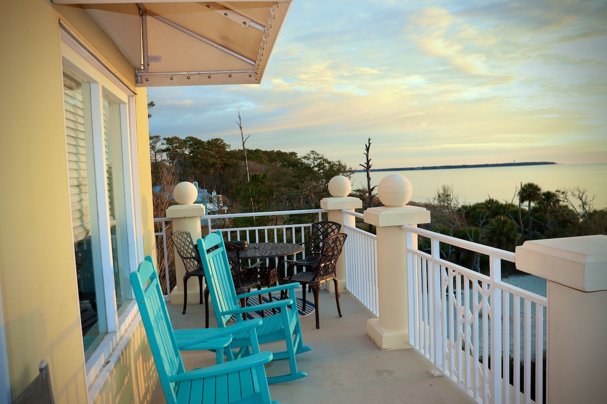 A wrap-around porch features turquoise chairs and a round table, providing a comfortable space for relaxation. The view extends towards the serene landscape and the horizon, where the sky transitions from soft pastel hues to the calm waters beyond.