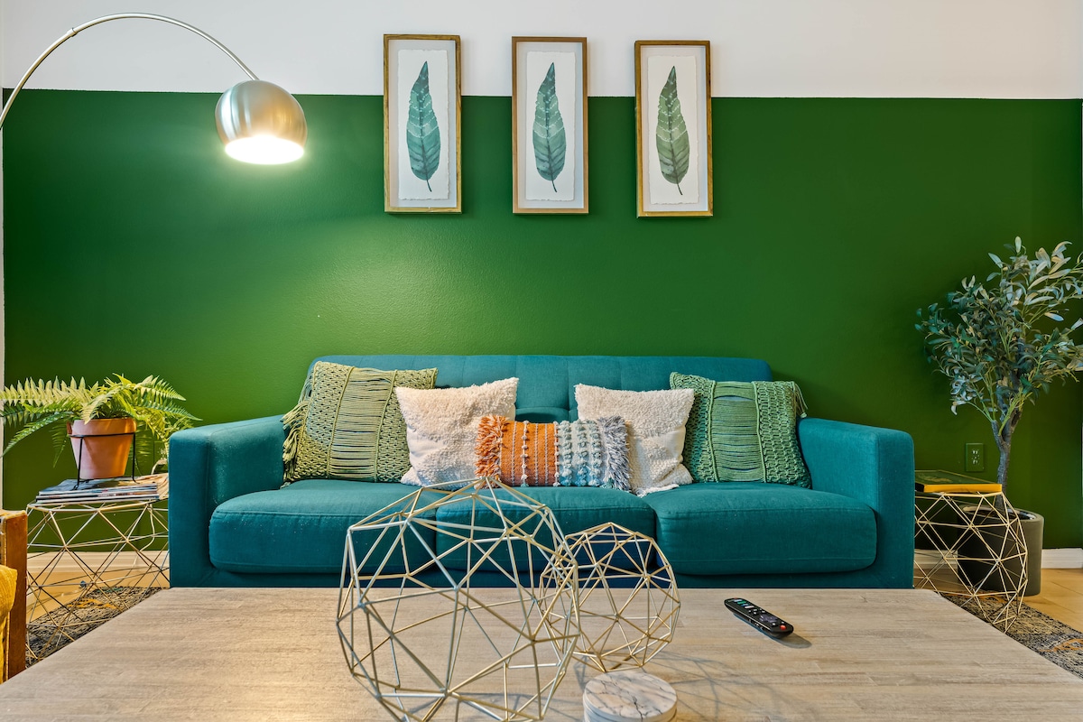 A vibrant living area features a teal sofa adorned with decorative pillows. A green accent wall enhances the space, while framed leaf prints add a touch of nature. A coffee table is positioned centrally, decorated with geometric wire sculptures and a remote control.