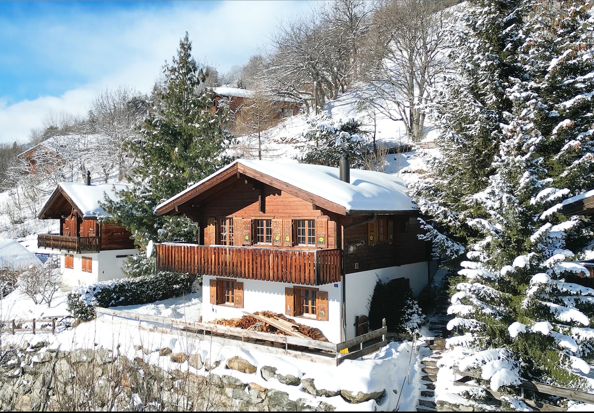 A charming chalet is seen nestled in a snowy landscape, featuring traditional wooden architecture. The sloped roof is blanketed in snow, while the balcony offers views of the surrounding winter scenery. Evergreen trees surround the property, enhancing the serene atmosphere of the location.