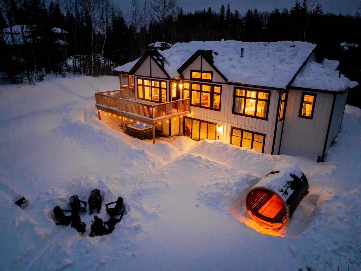 The exterior of the lakefront home is illuminated in warm light against a snowy landscape. The deck features ample seating and a sauna, while an outdoor seating area with Adirondack chairs surrounds a glowing fire pit, creating a welcoming space for gatherings.