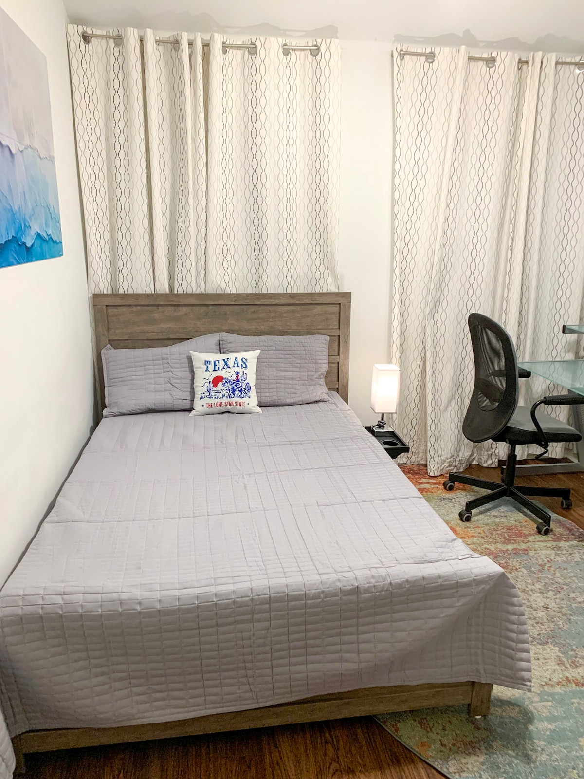 A cozy room features a comfortable bed with a light grey quilt and a decorative pillow displaying 'Texas.' Nearby, a modern desk and ergonomic chair provide a workspace. Soft lighting is offered by a standing lamp, with long, patterned curtains framing the window.