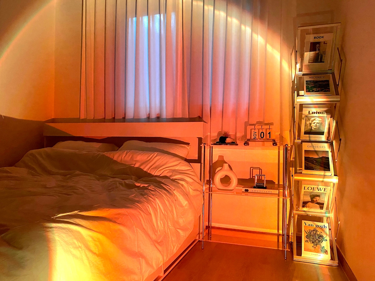 A cozy sleeping area features a spacious bed with soft bedding, illuminated by warm natural light filtering through sheer curtains. A decorative shelf holds various books, while a small round object and a calendar add an inviting touch to the space.
