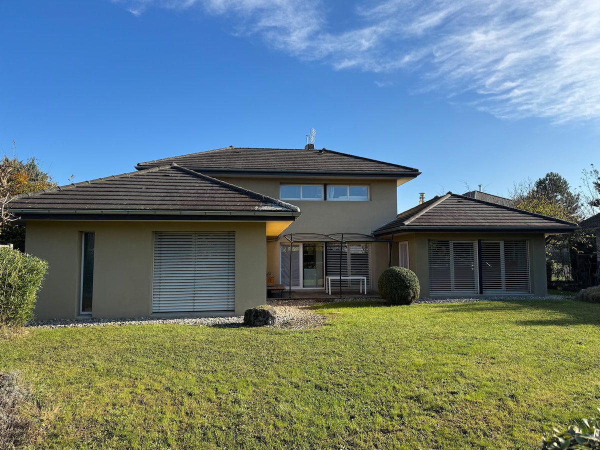 The exterior of a modern villa is depicted, featuring a spacious layout under a clear blue sky. Large windows dominate the façade, allowing for ample natural light. Lush green grass surrounds the building, enhancing the tranquil setting.