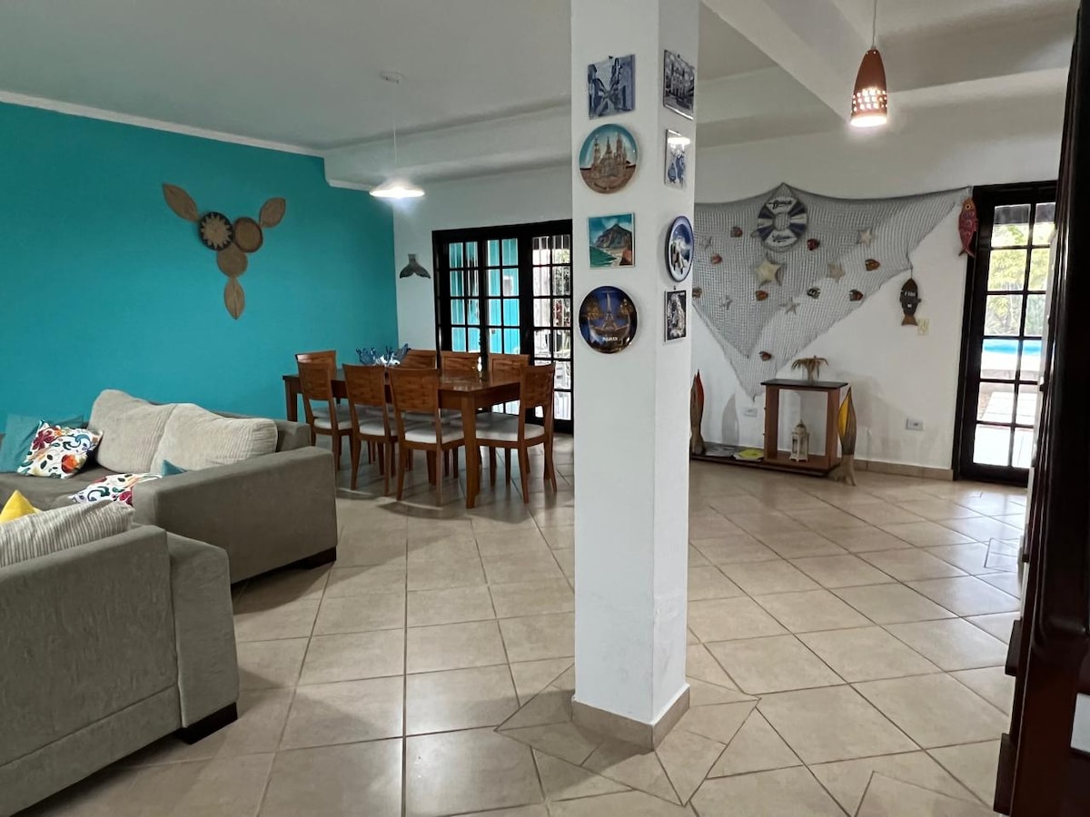An open living and dining area is presented, featuring turquoise walls and neutral tiled flooring. A spacious sofa and a large dining table with multiple wooden chairs are visible. Decorative wall art adds character, while large glass doors provide views of the outdoor pool area.