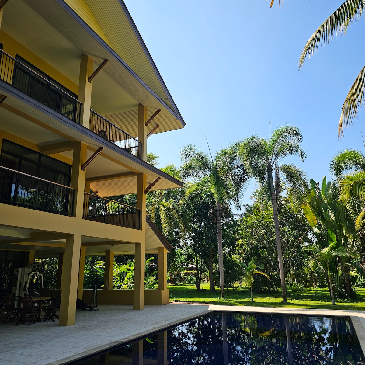A modern, multi-level building is depicted, featuring large balconies that overlook a serene pool. Lush greenery surrounds the area, with palm trees adding to the tropical landscape. Clear blue skies enhance the tranquil atmosphere, inviting relaxation and enjoyment.